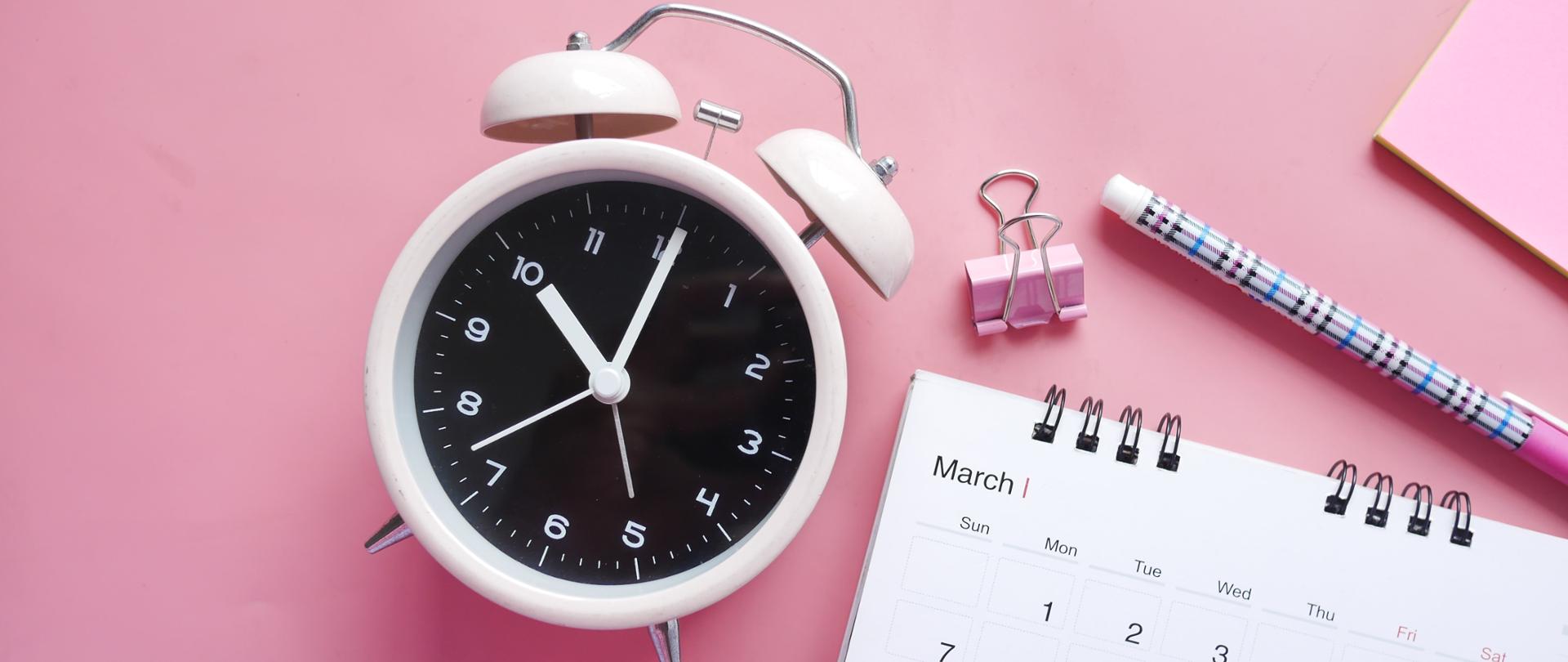 fotografia przedstawiająca jasnoróżowy budzik, różowy klips do papieru, długopis oraz fragment kalendarza oraz kolorowej kartki, całość zamieszczona na różowym tle