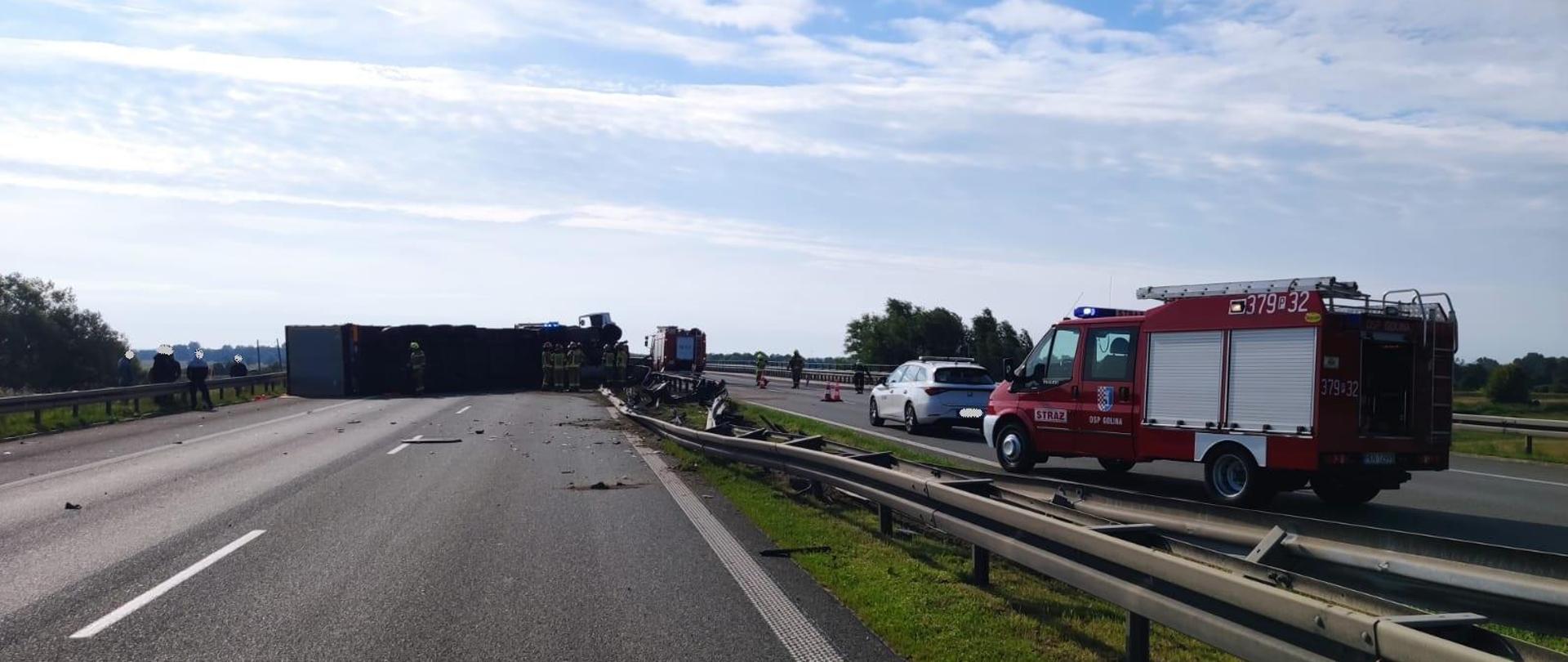Zdjęcie przedstawia przewrócony samochód ciężarowy wraz z naczepą na jezdnię Autostrady A2. Na zdjęciu widać również strażaków, samochody strażackie oraz samochód utrzymania autostrady. Barierki ochronne zostały uszkodzone. 