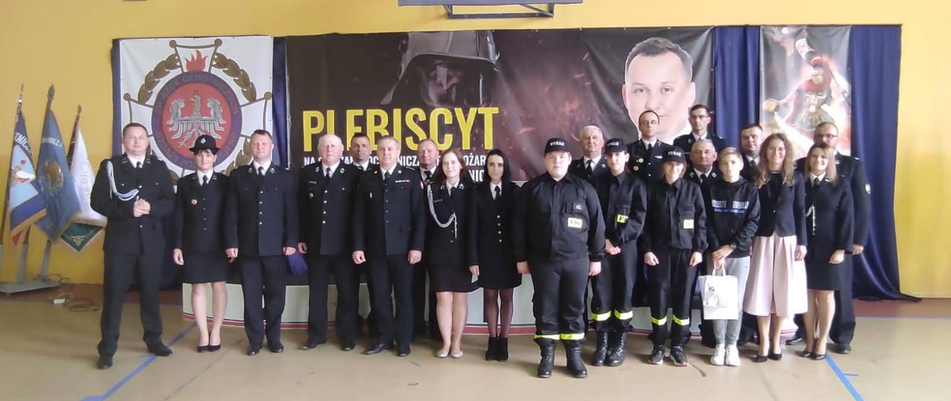 Na zdjęciu znajdują się strażacy z powiatu chełmińskiego oraz poseł Mariusz Kałużny podczas gali finałowej plebiscytu Strażak Roku 2021. Zdjęcie wykonano w sali sportowej Szkoły Podstawowej w Rogowie.
