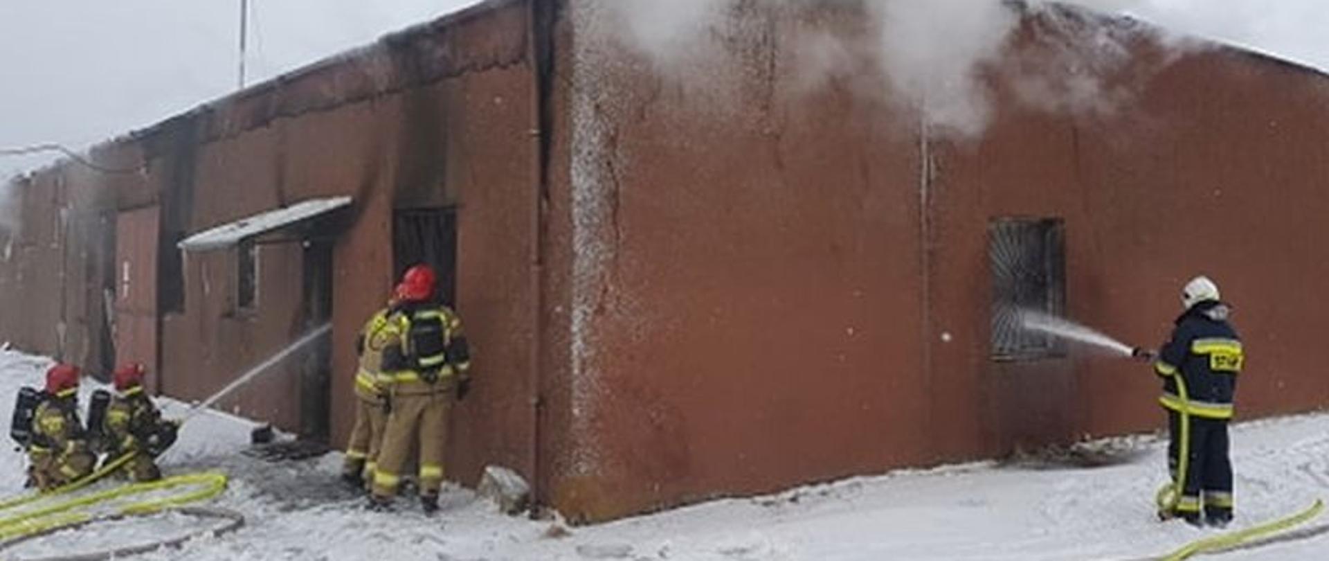 Okopcony budynek murowany, przez okna strażacy prowadzą akcję gaśniczą – podają wodę, nad budynkiem unosi się dym.