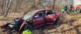 Zdjęcie przedstawia rozbity pojazd osobowy marki Skoda Oktavia. W tle dwa wozy bojowe straży pożarnej i dwóch strażaków.
