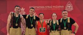 Pięciu uczestników wydarzenia sportowego pozuje do wspólnego zdjęcia w przestrzeni centrum handlowego. Cztery osoby mają na sobie spodnie strażackie oraz koszulki, a jedna osoba jest ubrana w strój sportowy i ma na szyi medal. W tle znajduje się czerwony baner Państwowej Straży Pożarnej z jej oznaczeniami.