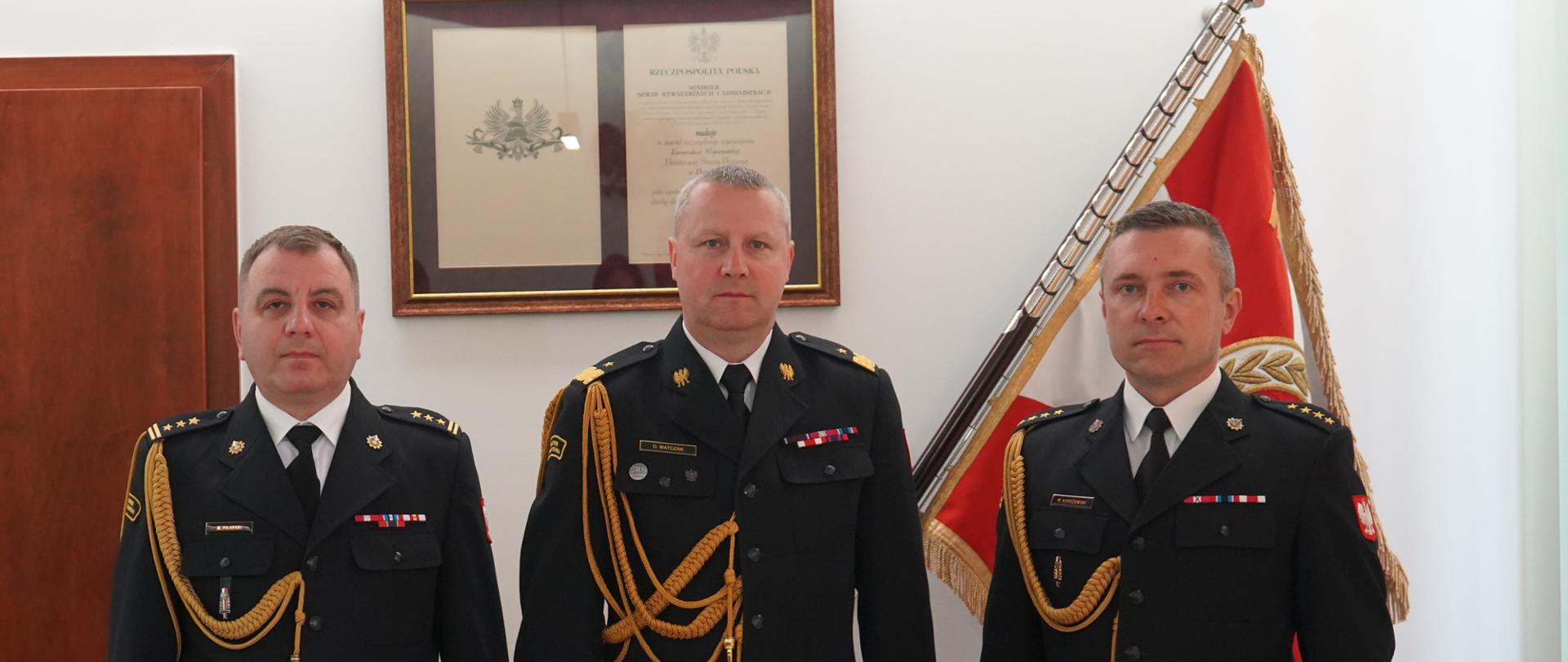 trzech strażaków w ciemnych mundurach ze sznurem stoi obok siebie, w tle sztandar