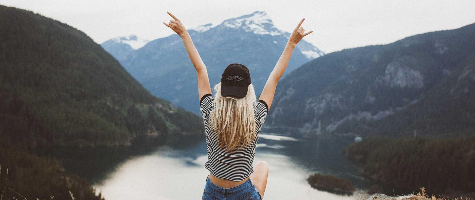 kobieta z uniesionymi rękoma siedząca na tle jeziora i gór
