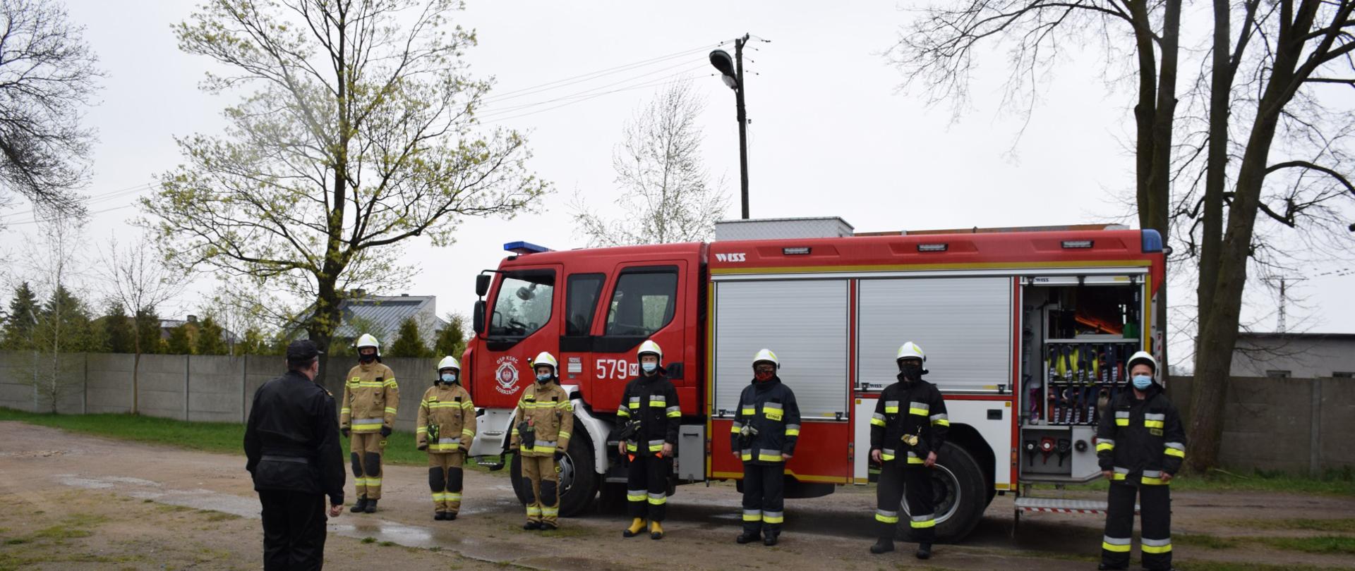 Strażacy w ubraniach specjalnych i maseczkach higienicznych, stoją w szeregu przy zachowaniu 1,5 m odległości. Z tyłu samochód pożarniczy.