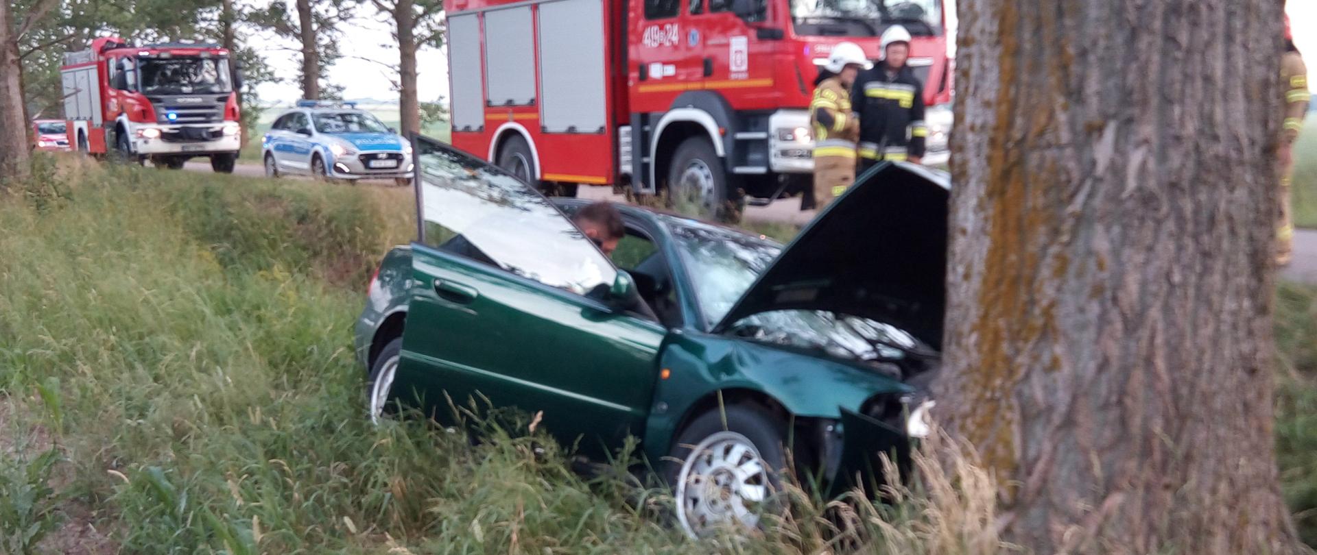 Samochód osobowy po uderzeniu w drzewo. Przód pojazdu zniszczony. Samochód stoi w przydrożnym rowie. W tle samochody strażackie i radiowóz