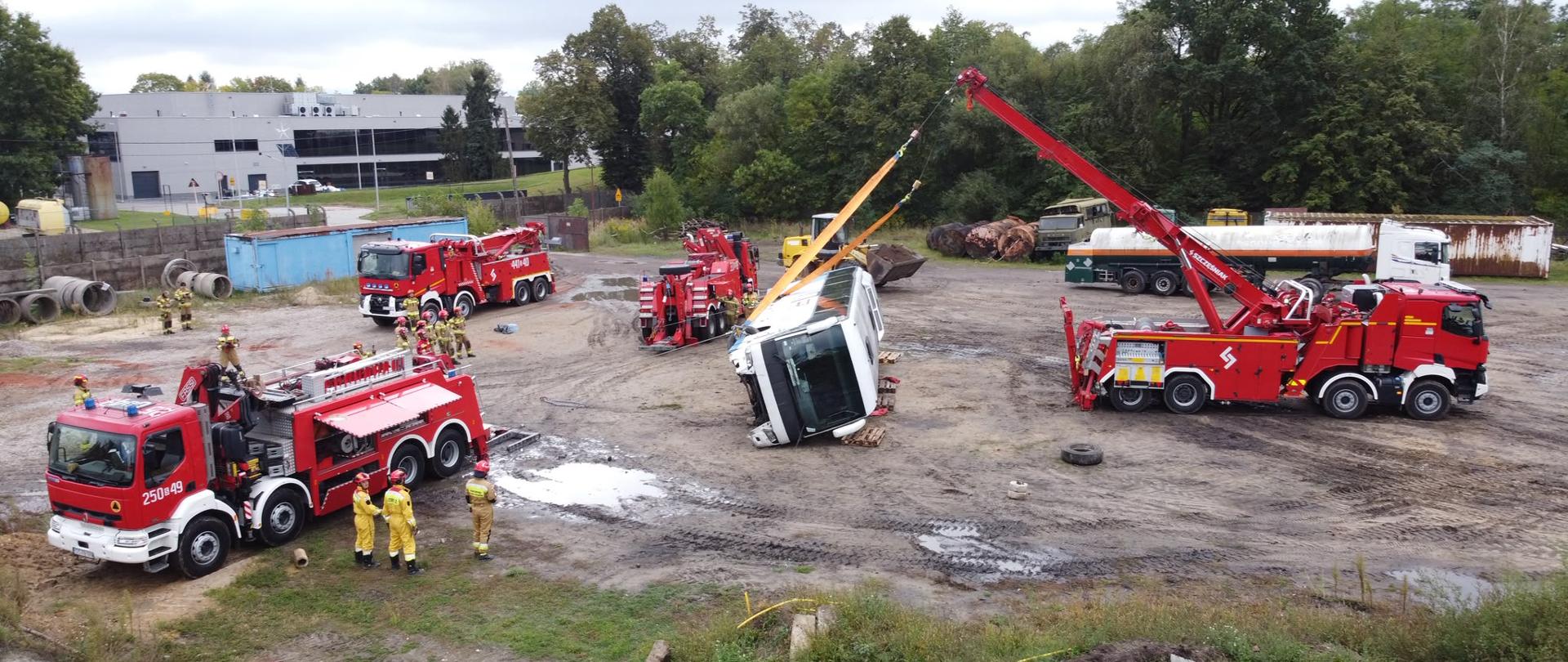 Poligon szkolny podczas ćwiczeń, na środku poligonu autobus przewrócony na bok który jest przymocowany linami przez samochód bojowy typy dźwig. Wokół autobusu stoją trzy inne samochody bojowe typu dźwig, w głębi po prawej stoi samochód typu cysterna