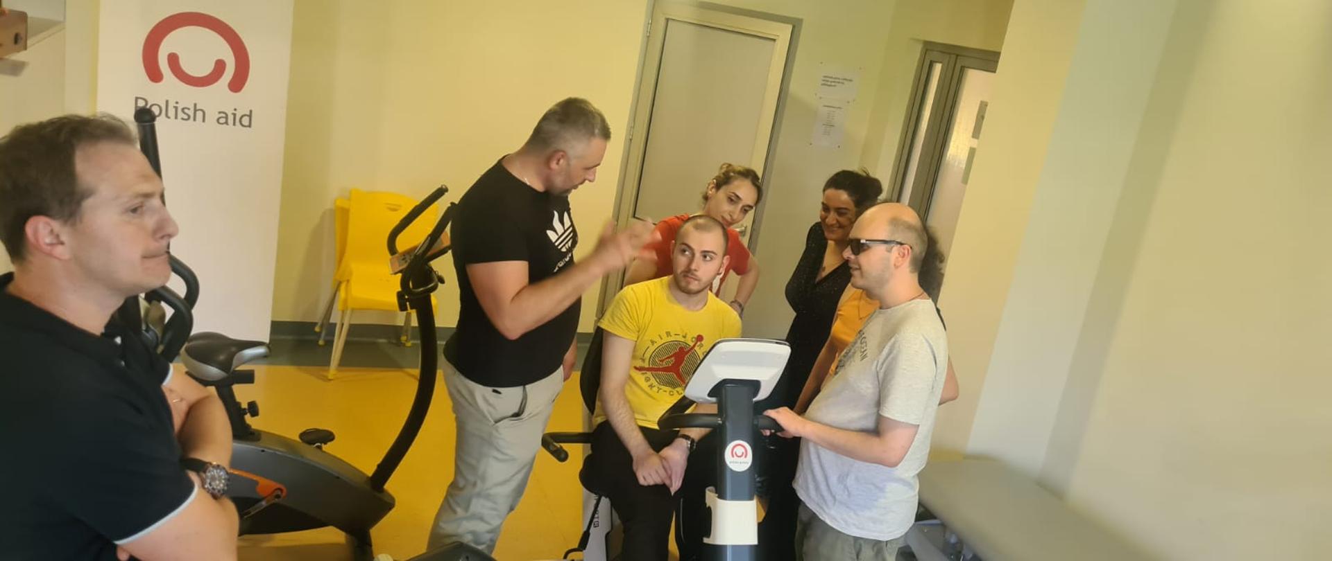 Jedna osoba siedzi na rowerku treningowym z naklejką loga Polska pomoc, 6 innych osób stoi wokół niej i rozmawia, z tyłu widać banner z logo Polish aid