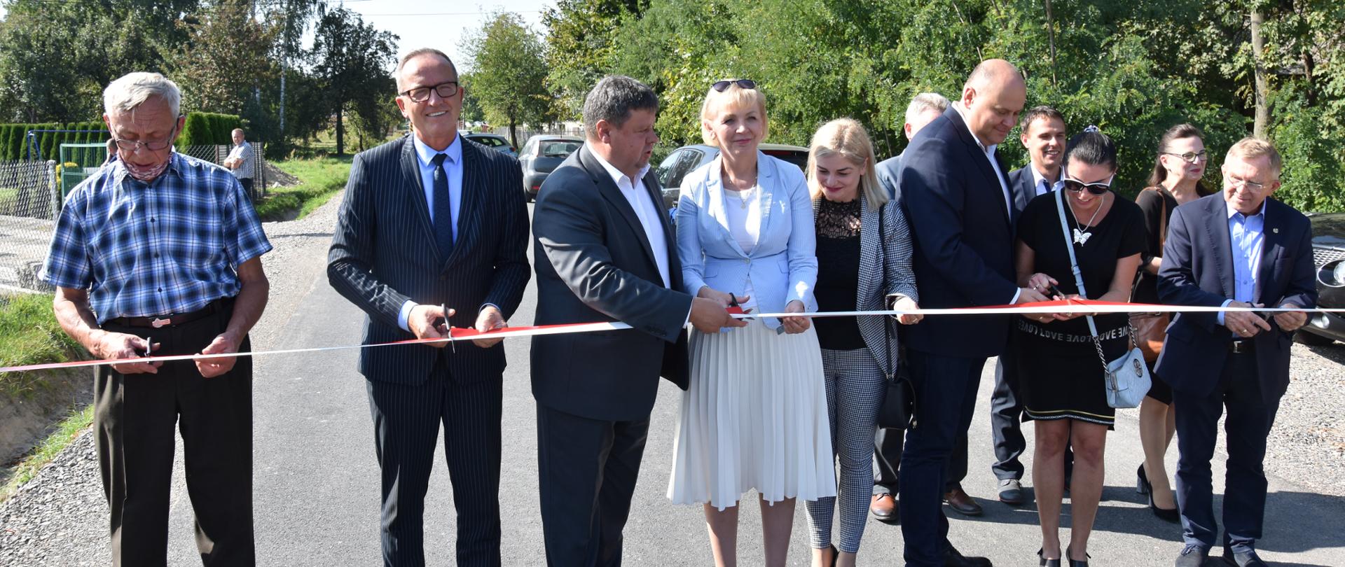 Zmodernizowane drogi oddano do użytku poprzez symboliczne przecięcie biało-czerwonej wstęgi. Na pierwszym planie znajdują się elegancko ubrane osoby przecinający wstęgę. Osoby te stoją na zmodernizowanej drodze
