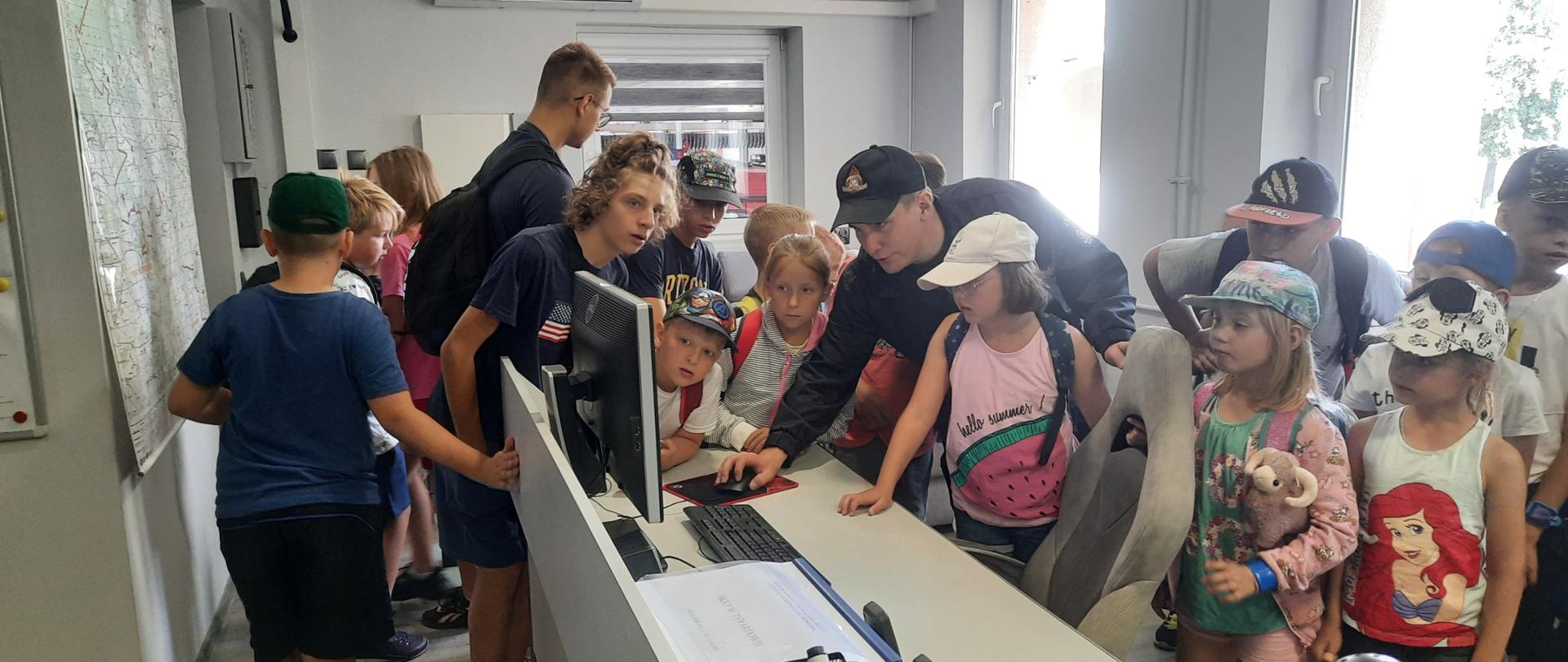 Uczestnicy półkolonii stoją przy pulpicie stanowiska kierowania, strażak pokazuje im jak przyjmuje się zgłoszenie na komputerze