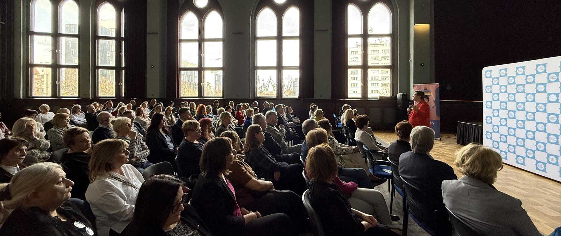 Duża aula, na krzesłach ustawionych w rzędach siedzi duża grupa osób, przed nimi z mikrofonem stoi wiceminister Katarzyna Lubnauer 