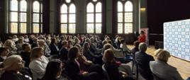 Duża aula, na krzesłach ustawionych w rzędach siedzi duża grupa osób, przed nimi z mikrofonem stoi wiceminister Katarzyna Lubnauer 