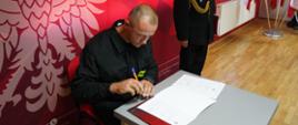 Świetlica komendy. Na tle ścianki promocyjnej przy stoliku siedzi strażak, który podpisuje akt ślubowania. Ma na sobie ubranie koszarowe w kolorze ciemnym. Obok stoi zastępca komendanta powiatowego w umundurowaniu wyjściowym ze sznurem galowym. W tle widoczny jest stojak z flagami biało-czerwonymi.