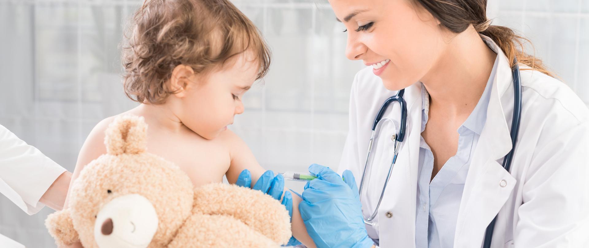 Na zdjęciu młoda uśmiechnięta lekarka szczepi małe dziecko, które trzyma pluszowego misia.