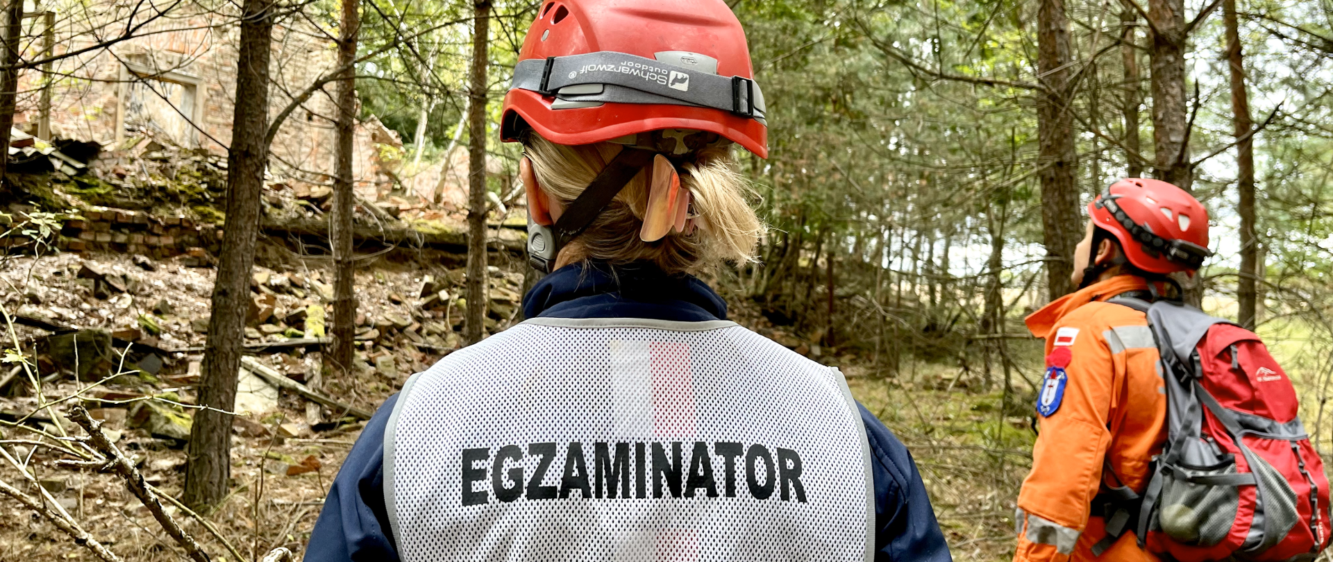 Dwie postaci stojące tyłem. Obie mają na głowach czerwone kaski, jedna z nich na plecach ma napis EGZAMINATOR, druga posiada plecach. Obie są w otoczeniu drzew – lasu. 