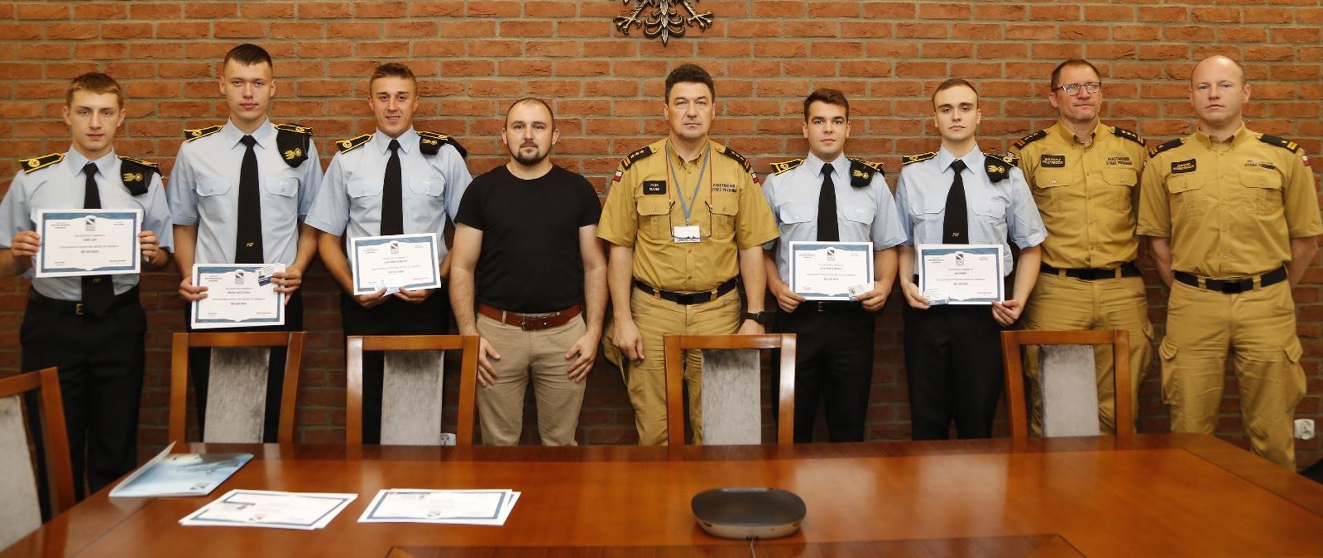 Grupowe zdjęcie uczestników szkolenia (pięciu kadetów CS PSP) w towarzystwie Komendanta Centralnej Szkoły PSP, instruktora oraz dwóch oficerów CS PSP na tle ściany z wiszącym metalowym godłem RP