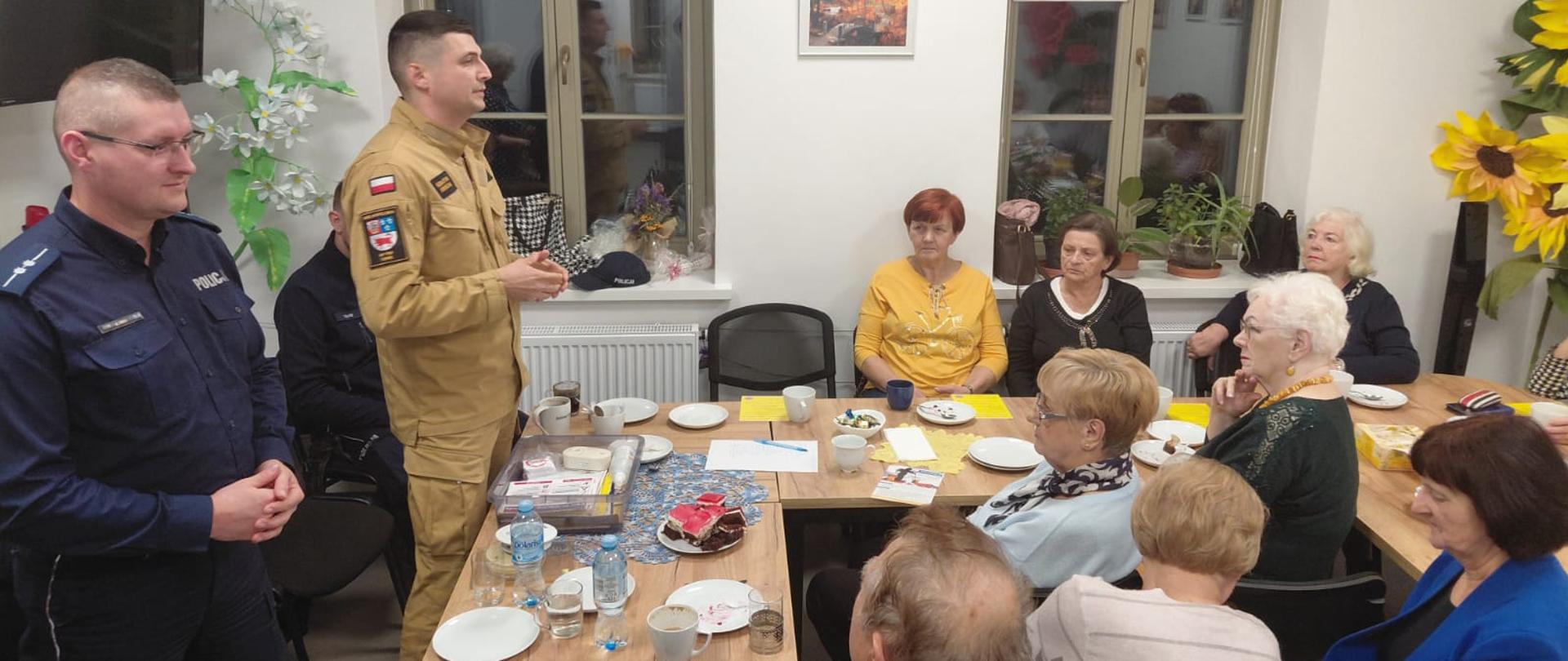 Grupa starszych osób siedzi przy stołach w jasnym pomieszczeniu, słuchając dwóch umundurowanych funkcjonariuszy – jednego w granatowym stroju policyjnym, drugiego w beżowym mundurze. Na stołach stoją talerze, kubki i drobne przekąski, a w tle widać okna oraz dekoracje roślinne.