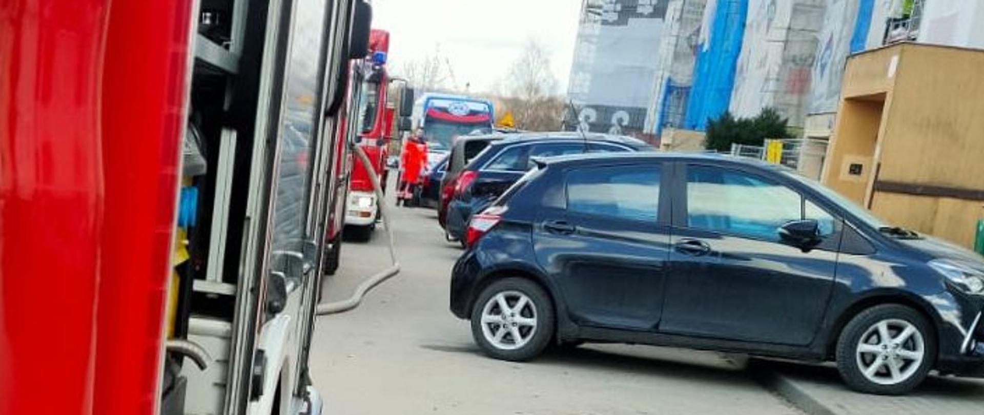 Po lewej stronie stoją w rzędzie wzdłuż ulicy pojazdy pożarnicze, po prawej stronie znajduje się blok mieszkalny. W centralnej części stoją zaparkowane przy chodniku auta osobowe.