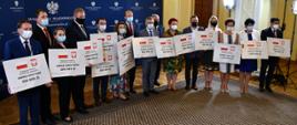 Na zdjęciu znajduje się 15 elegancko ubranych osób. Większość z nich trzyma w rękach symboliczne promesy ze środkami wsparcia finansowego z Rządowego Funduszu Inwestycji Lokalnych. 