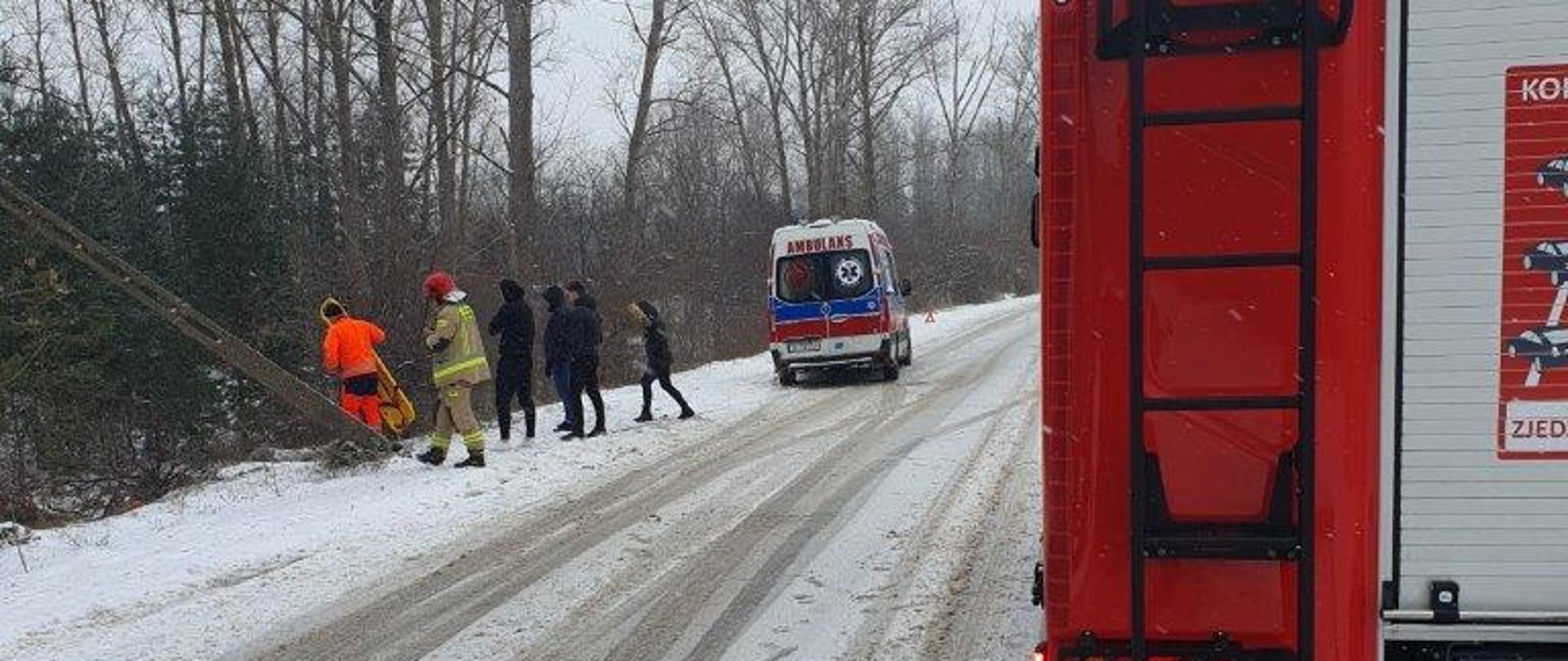Zdjęcie przedstawia ulicę Chorzowską w Kielcach. Po prawej stronie widać fragment samochodu pożarniczego, skrajnie po lewej widać ratowników schodzących przy pochylonym betonowym słupie oświetlenia ulicznego do rowu gdzie leży przewrócony samochód osobowy. W głębi widać ambulans Państwowego Ratownictwa Medycznego.