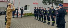 Zdjęcie przedstawia strażaków stojących na palcu przed budynkiem strażnicy podczas uroczystego apelu z okazji Dnia Flagi. 
