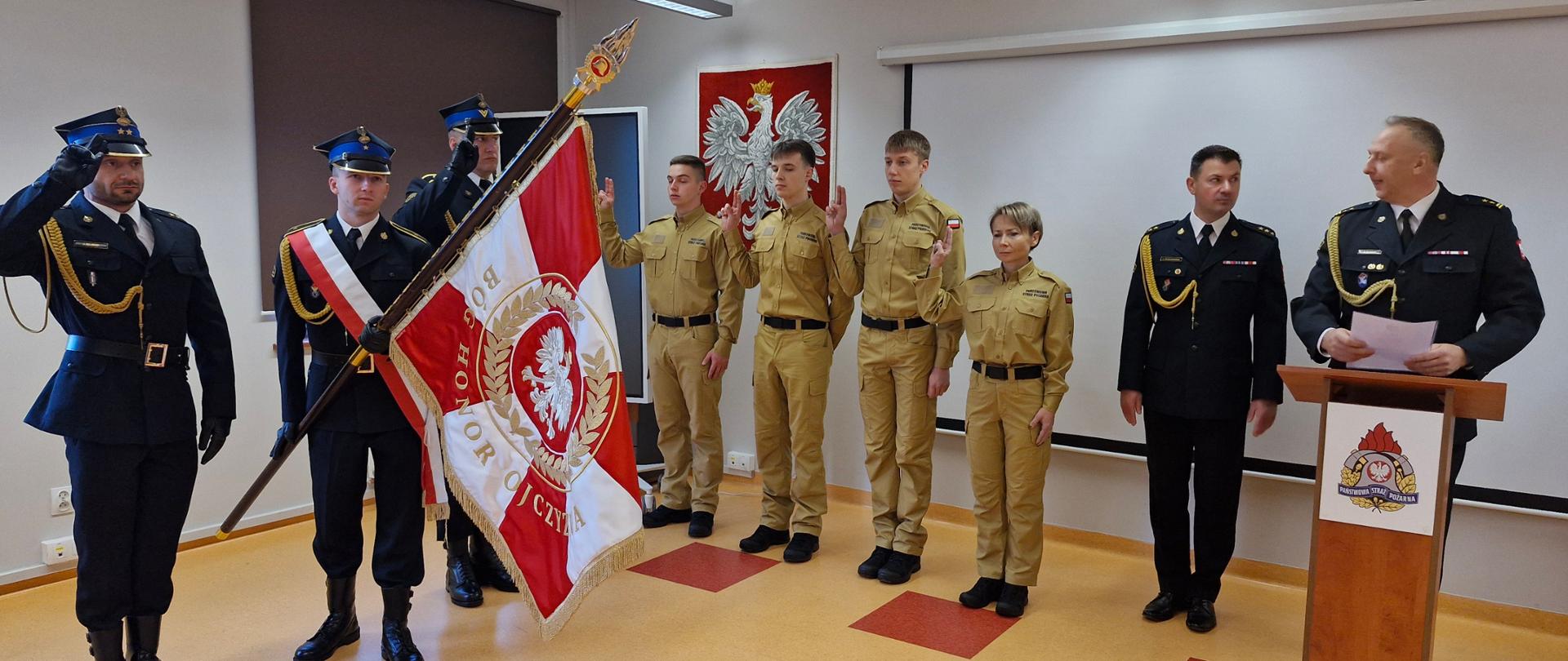 Moment ślubowania – osoby w beżowych mundurach stoją z uniesioną prawą ręką. Obok nich strażacy w galowych mundurach oraz osoba trzymająca sztandar z napisem „Bóg, Honor, Ojczyzna”