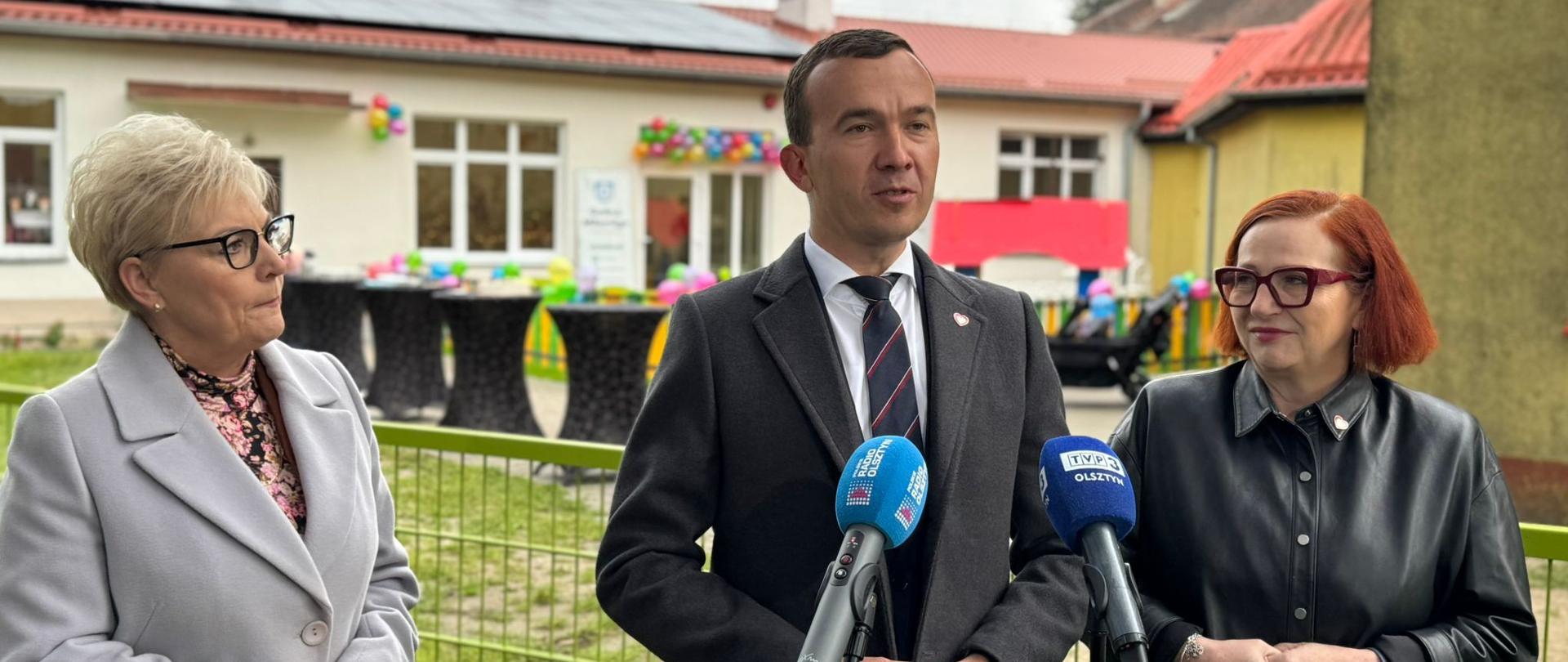 Mężczyzna stoi z dwiema kobietami i przemawia do mikrofonu 