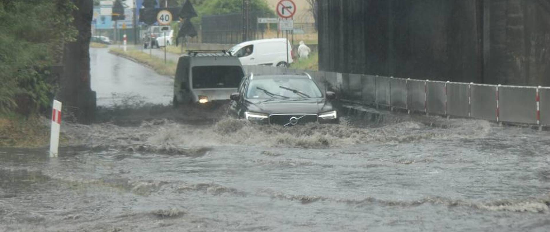 Zalany odciek drogi krajowej przebiegający pod wiaduktem kolejowym. Samochód osobowy przejeżdża przed zalany odcinek. Lustro wody sięga do świateł terenowego samochodu. 