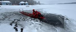Strażacy w czerwonych ubraniach wypornościowych do pracy w wodzie ćwiczą podejmowanie osoby poszkodowanej, pod którą załamał się lód i wpadła do wody. Na zdjęciu strażacy na saniach lodowych. Strażak podejmuje osobę poszkodowaną z wody, osoba poszkodowana przytomna.