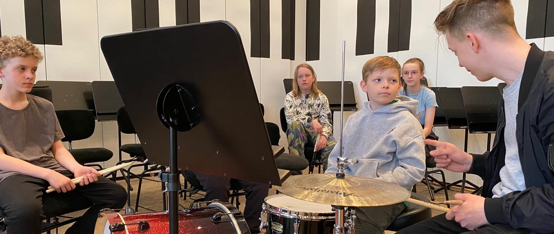 Mężczyzna siedzi z pałeczkami w rękach przy zestawie perkusyjnym, obok niego chłopiec, wokół nich z tyłu siedzi dwóch chłopców i dziewczyna na scenie auli PSM.
