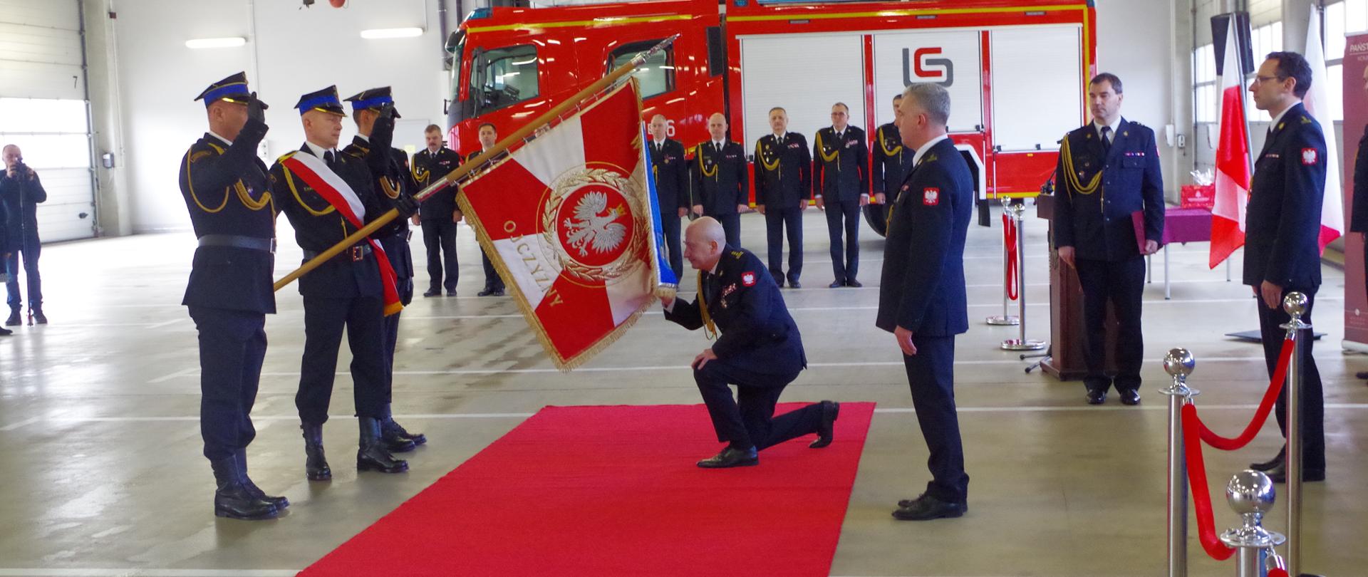 Na garażu Komendy Miejskiej PSP w Mysłowicach w obecności strażaków zdający obowiązki komendanta klęka na czerwonym dywanie przed sztandarem Jednostki Ratowniczo- Gaśniczej PSP w Mysłowicach