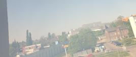 Dym wydobywa się z okna ZUS. W tle widać zabudowę miasta Kalisza. Wokół krawędzi zdjęcia rama okna budynku ZUS. Niebo błękitne, pogoda słoneczna.