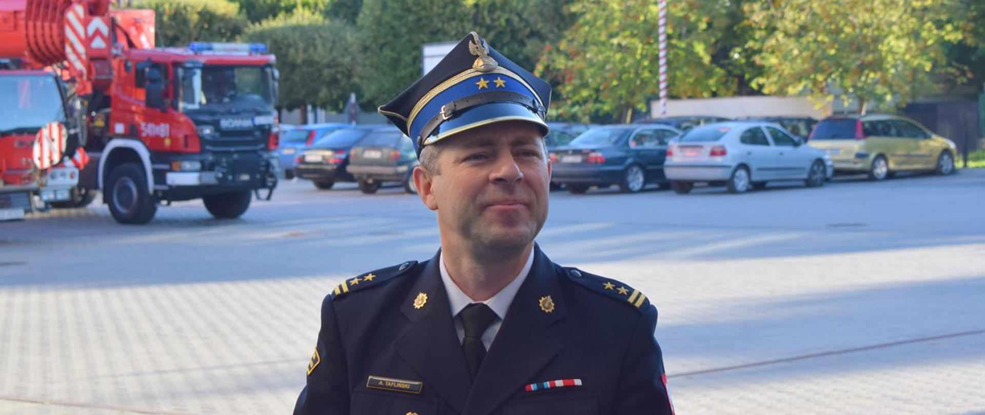 Wizerunek bryg. Adama Taflińskiego - w tle pojazdy pożarnicze i parking JRG z samochodami osobowymi