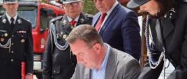 Goście wpisują się do księgi pamiątkowej Komendy Powiatowej PSP w Łowiczu, w tle stoją posłowie na sejm III RP oraz prezesa zarządu powiatowego OSP