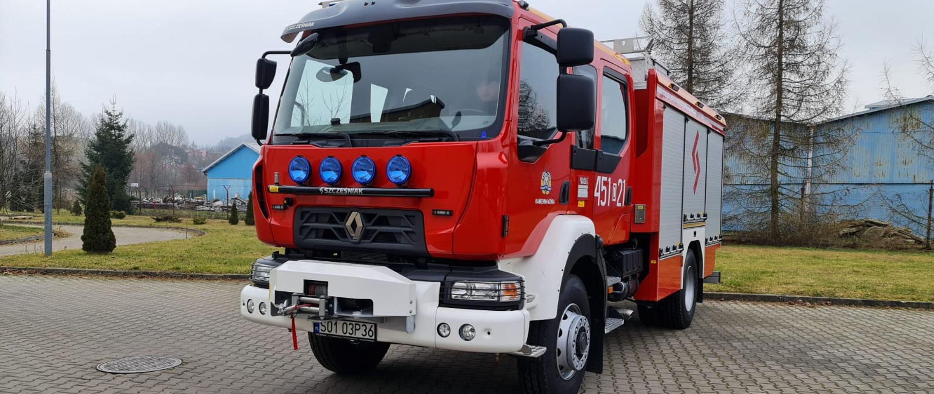 Samochód średni ratowniczo gaśniczy w kolorze czerwonym ustawiony lewym bokiem, widoczny przód pojazdu. Z boku widoczne oznaczenie 451 - 21. zaparkowane na betonowej kostce w tle widoczny teren zielony - trawa