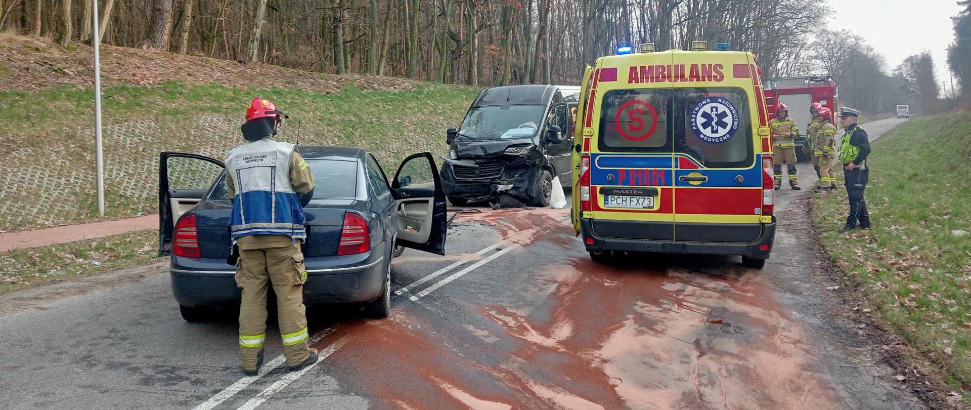 Zdjęcie przedstawia strażaków i policjantów działających przy wypadku drogowym.
W tle pojazdy uszkodzone, samochody strażackie i las.
