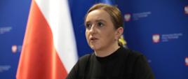 Wiceminister Olga Semeniuk wzięła udział w VII Kongresie Klastrów Polskich. Podczas Kongresu wiceminister siedzi przy stole. W tle baner resortu rozwoju, a po lewej stronie flaga Polski.