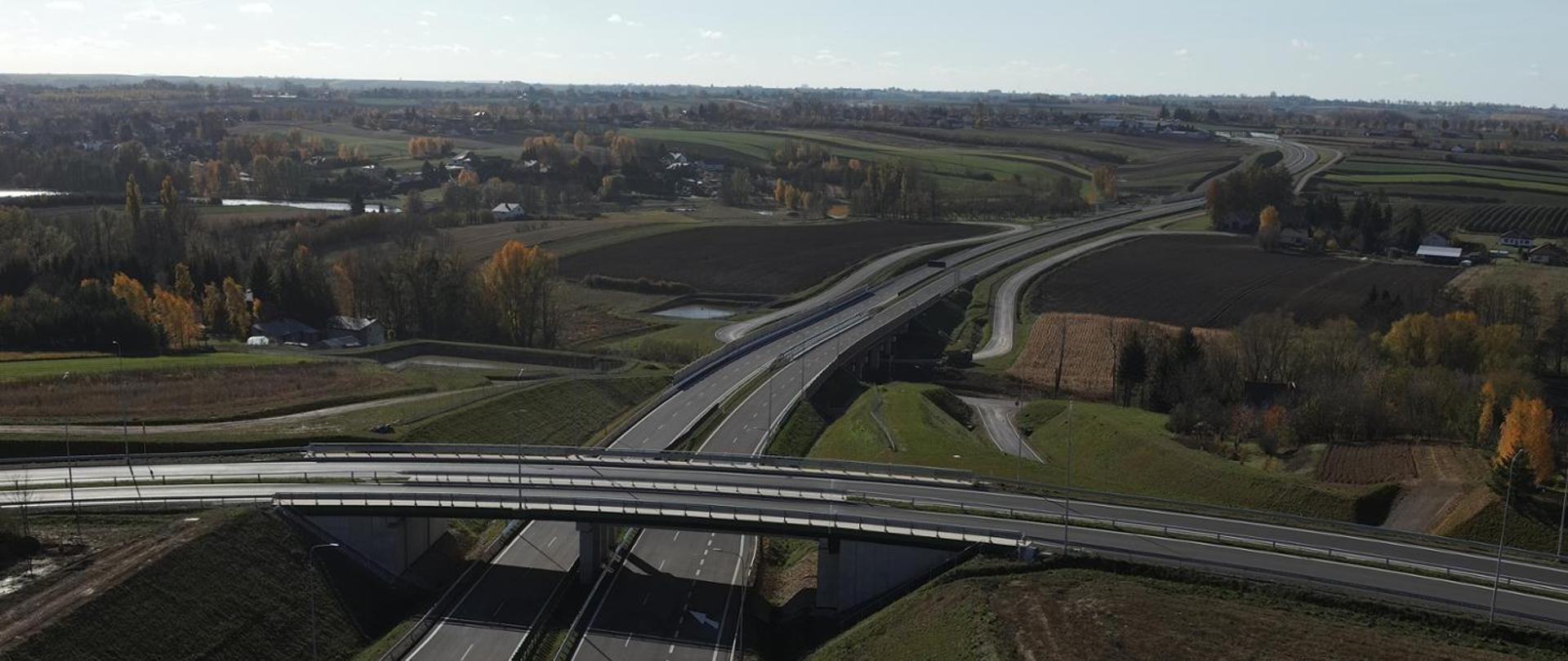 Widok z lotu ptaka na węzeł drogowy obwodnicy Opatowa, z kilkoma pasami ruchu i estakadą przecinającą trasę. W tle rozciągają się pola uprawne, zabudowania oraz jesienne drzewa.