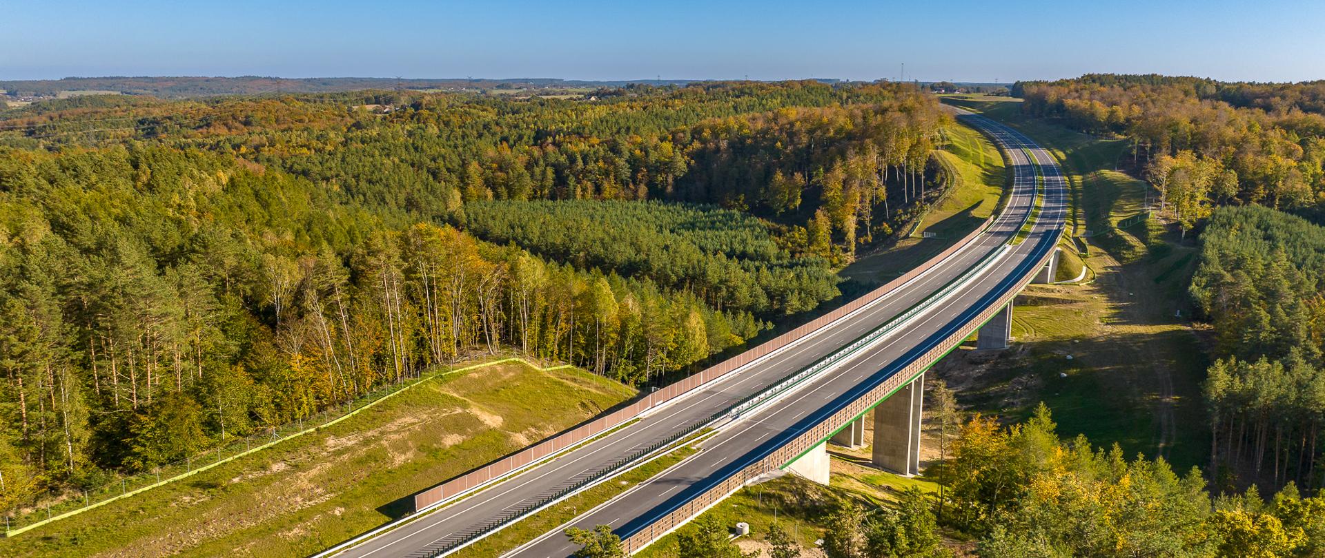 Zdjęcie przedstawia drogę ekspresową S6 na odcinku Luzino - Szemud. Widok z lotu ptaka ukazuje nowoczesną, wielopasmową drogę przecinającą malowniczy krajobraz pełen zieleni i lasów. Droga biegnie przez most, który wznosi się nad doliną, co dodaje zdjęciu dynamiki i ukazuje infrastrukturę wkomponowaną w naturalne otoczenie. Na horyzoncie widoczny jest rozległy obszar leśny, a niebo jest bezchmurne i jasnoniebieskie, co podkreśla piękno dnia.