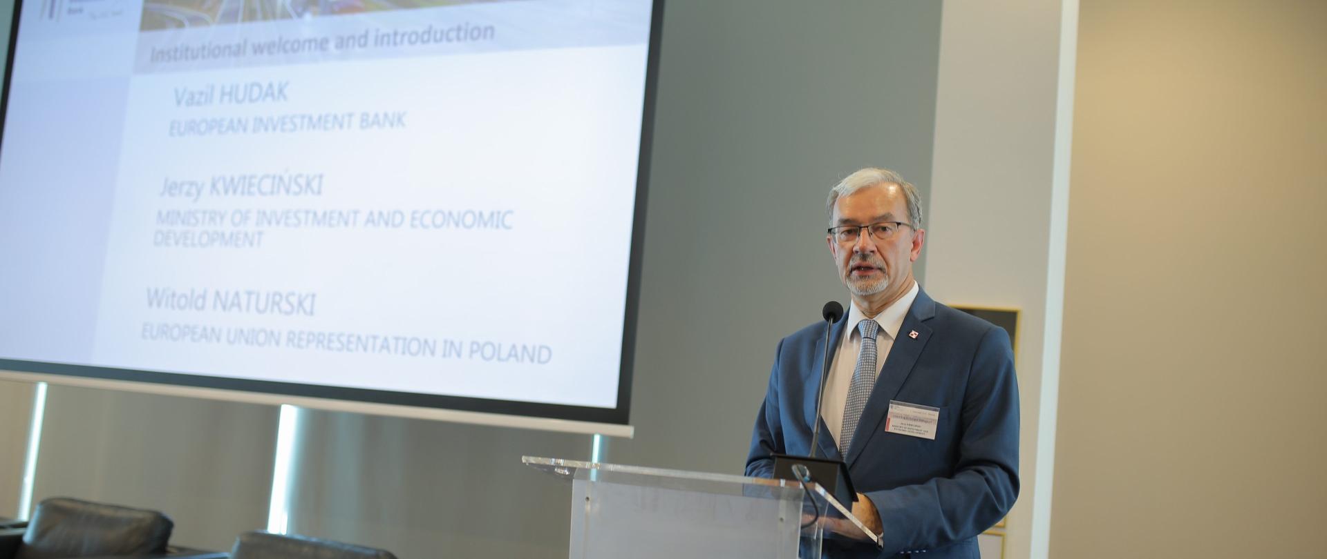Minister Jerzy Kwieciński stoi przy mównicy, za nim na tablicy wyświetlany jest napis z nazwiskami prelegentów, Vazil Huda, Jerzy Kwieciński, Witold Naturski. 