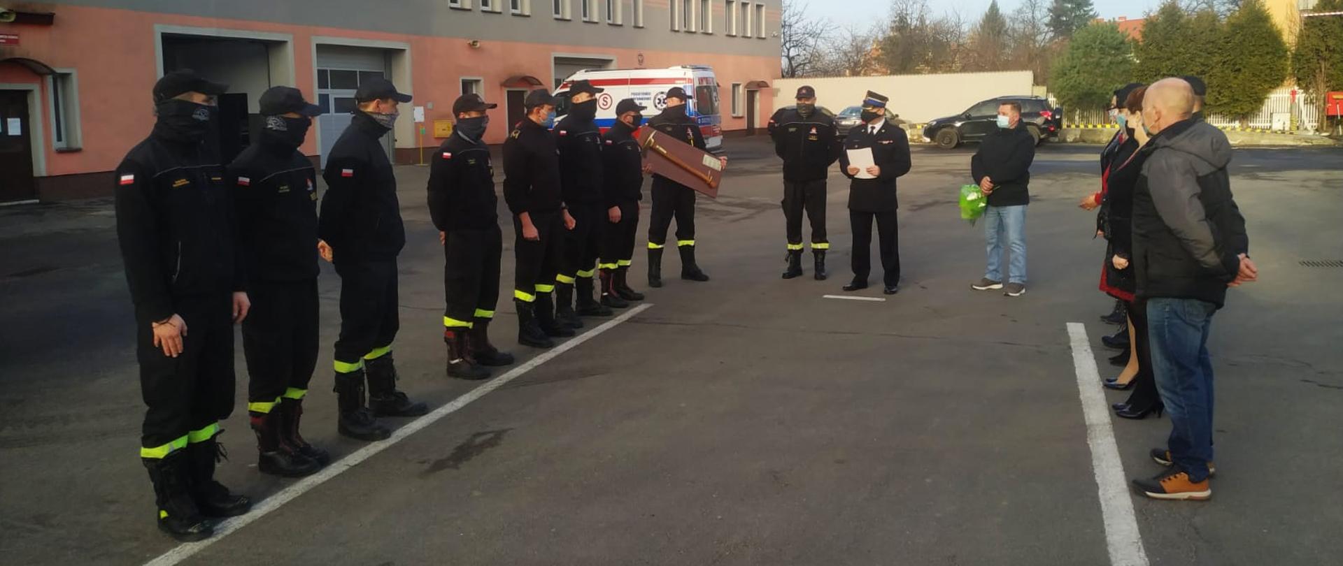 Obraz przedstawia strażaków ustawionych w dwóch szeregach naprzeciw siebie. W środkowej części Komendant Powiatowy brygadier Waldemar Sarlej dziękujący za służbę starszemu ogniomistrzowi Pawłowi Rozmusowi. W tle budynek komendy powiatowej.