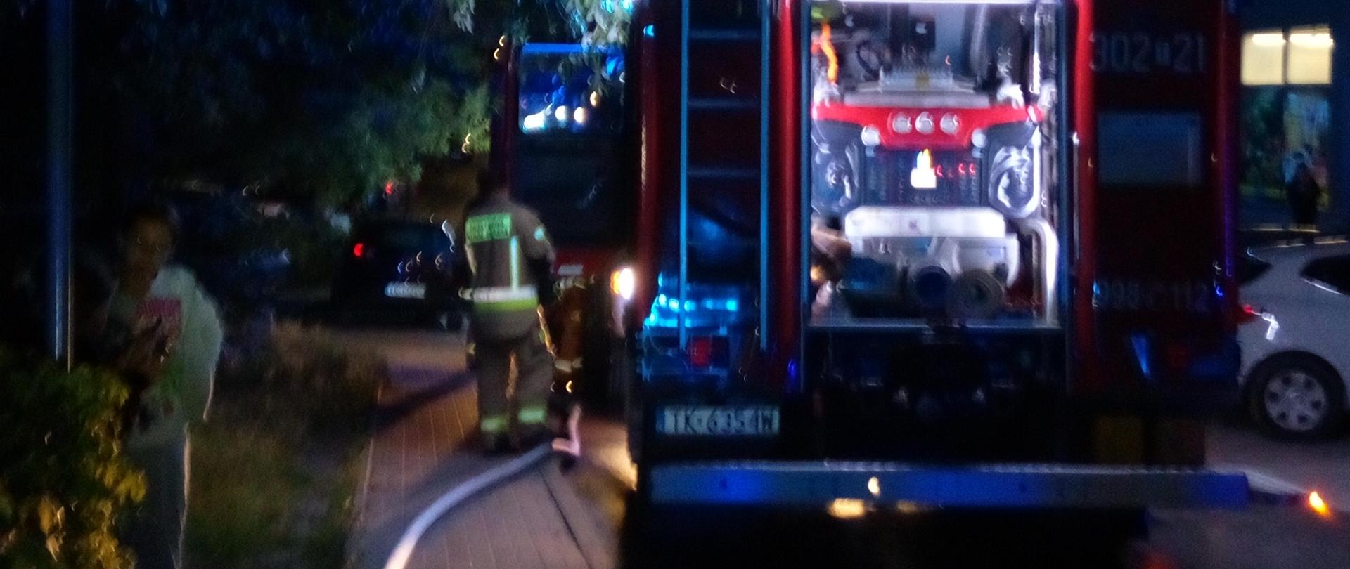 Zdjęcie przedstawia ulicę Wiosenną. Na jezdni zaparkował samochód pożarniczy. Chodnikiem poprowadzono wąż gaśniczy wypełniony wodą. Przy aucie stoi ratownik.