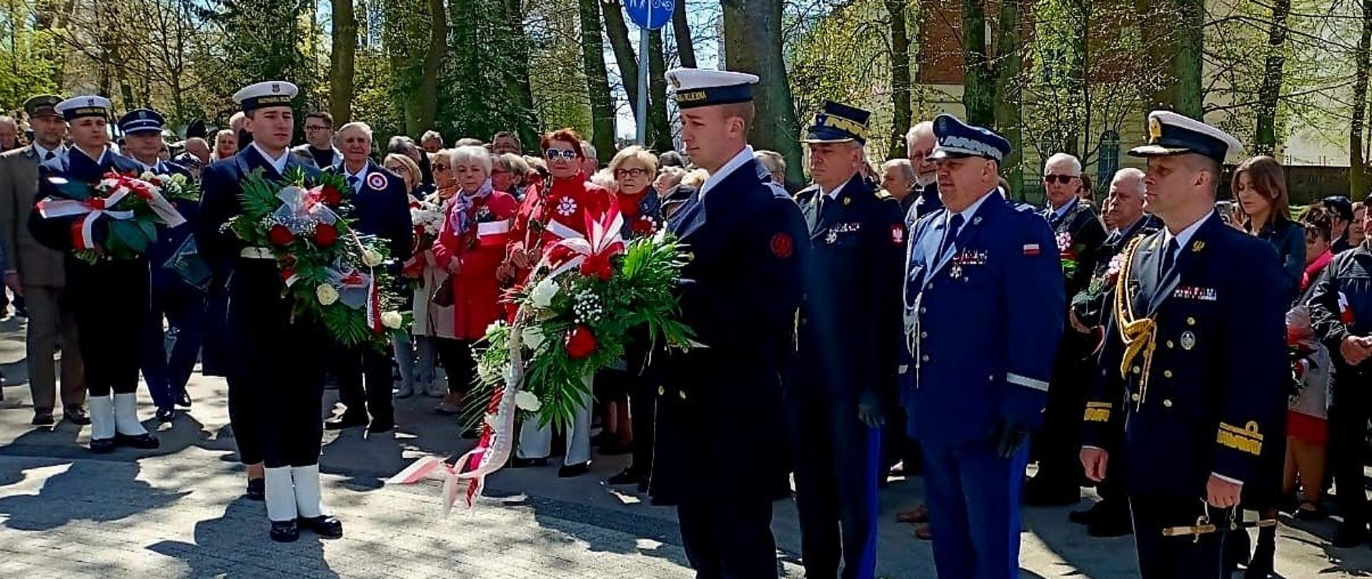 Przedstawiciele Państwowej Straży Pożarnej Policji oraz Straży Granicznej stoją za żołnierzem trzymającym wiązankę kwiatów. Obok stoją osoby biorące udział w uroczystości.