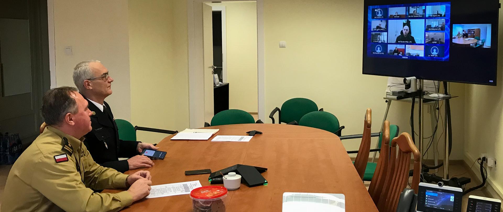 Na zdjęciu widać dwóch strażaków podczas wideokonferencji. Strażacy siedzą przy stole po prawej stronie fotografii. Z lewej strony u góry widać monitor telewizora. Po środku biurko z dokumentami.
