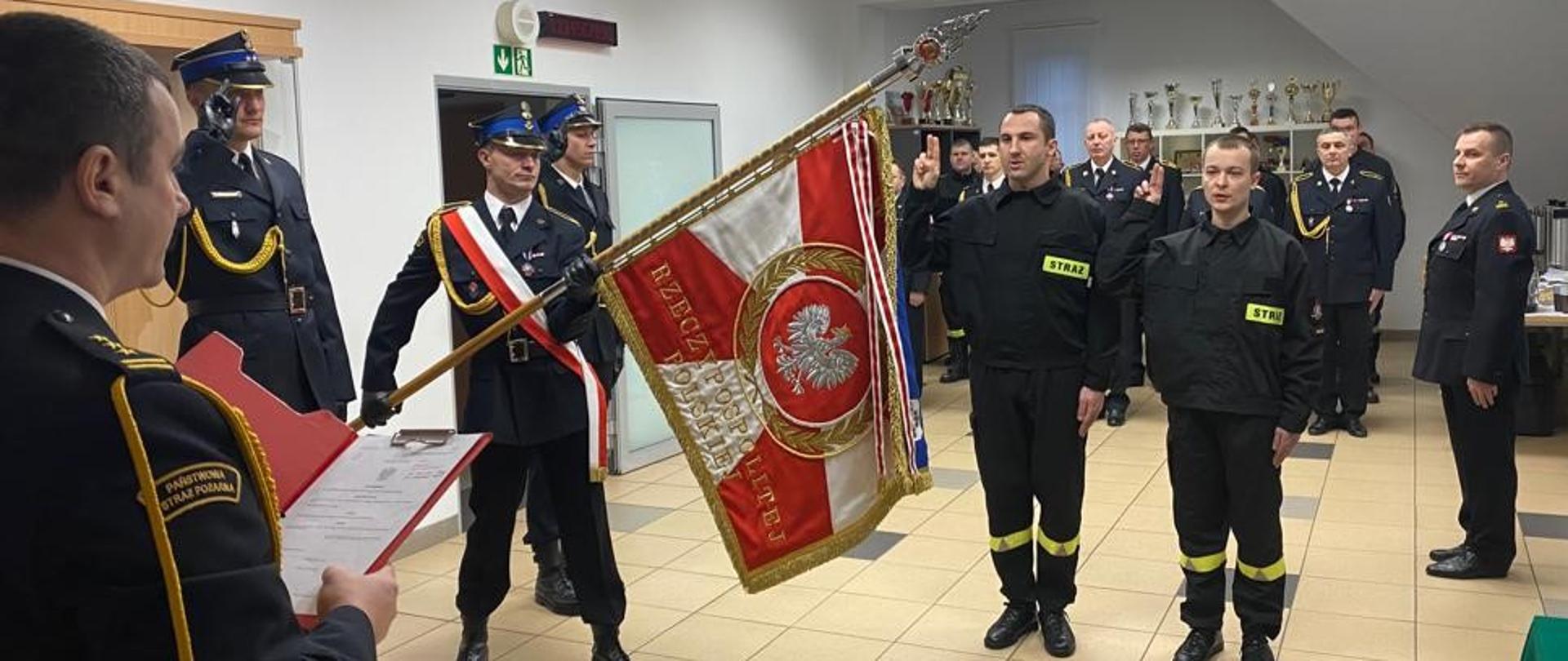 Zdjęcie przedstawia uroczysty moment składania ślubowania przez strażaków nowo przyjętych do służby w Komendzie Powiatowej Państwowej Straży Pożarnej w Gołdapi. Z lewej strony obrazu znajduje się Komendant Powiatowy Państwowej Straży Pożarnej w Gołdapi st. bryg. Wojciech Szczepanik, który przyjmuje ślubowanie oraz poczet sztandarowy. W tle można zaobserwować pozostałych strażaków biorących udział w uroczystej zbiórce