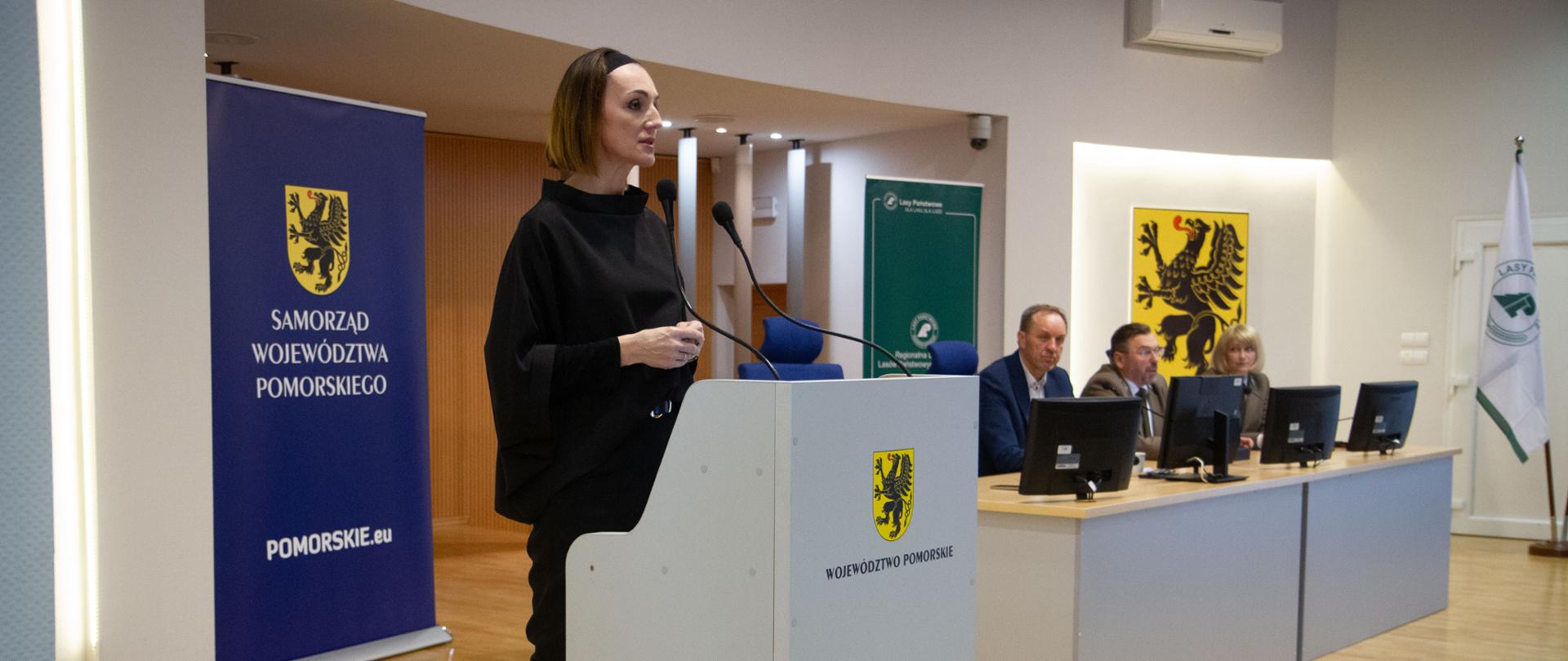 sala, kobieta (wojewoda pomorska) stoi za mównicą, obok za stołem prezydialnym trzech mężczyzn, jeden w mundurze leśnika