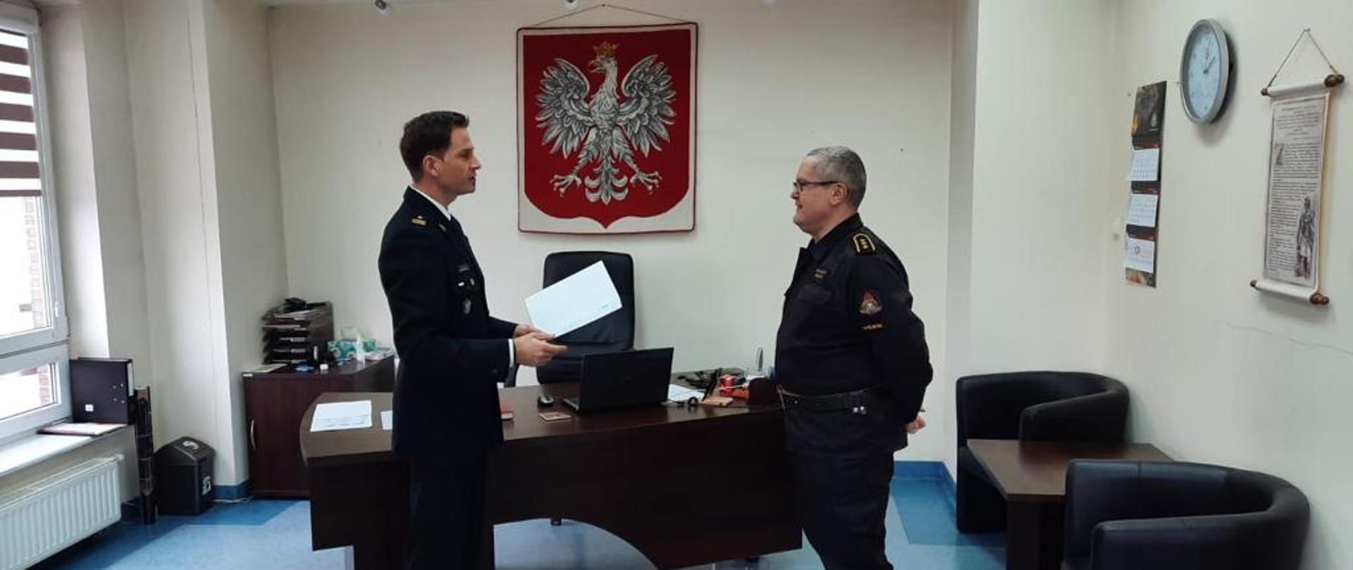 Na zdjęciu Komendant Powiatowy PSP w Myśliborzu na tle godła Rzeczposolitej Polskiej wręcza funkcjonariuszowi okolicznościowy dokument zawierający podziękowania. Obaj funkcjonariusze ubrani w umundurowanie służbowe.
