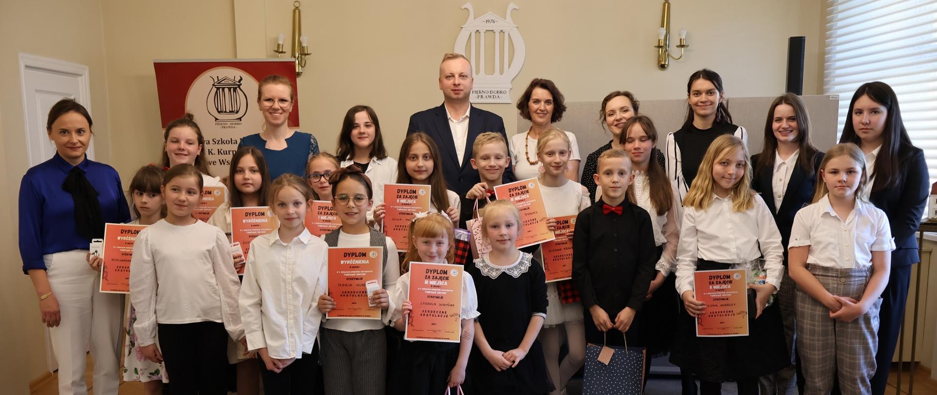 Uczniowie i nauczyciele stojący w auli szkoły.