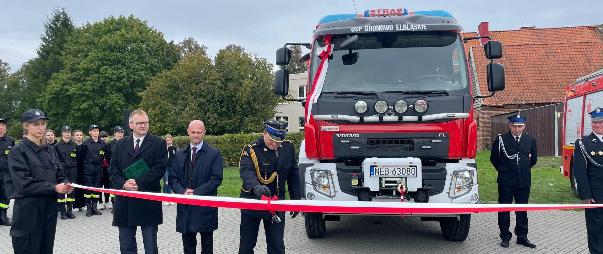 Strażacy w mundurach oraz zaproszeni goście stoją przed biało-czerwoną wstęgą i przesuwają po niej kluczyki, za nimi pojazd ratowniczo-gaśniczy. W tle zgormadzeni strażacy.