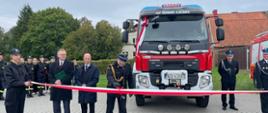 Strażacy w mundurach oraz zaproszeni goście stoją przed biało-czerwoną wstęgą i przesuwają po niej kluczyki, za nimi pojazd ratowniczo-gaśniczy. W tle zgormadzeni strażacy.